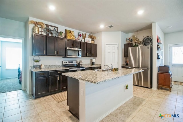 kitchen featuring sink, light tile patterned floors, stainless steel appliances, light stone counters, and an island with sink