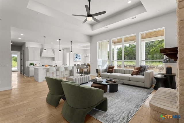 living room featuring ceiling fan with notable chandelier, light hardwood / wood-style floors, sink, and a tray ceiling