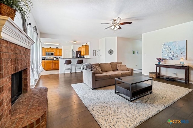 living area with dark wood-style floors, a ceiling fan, a brick fireplace, a textured ceiling, and baseboards