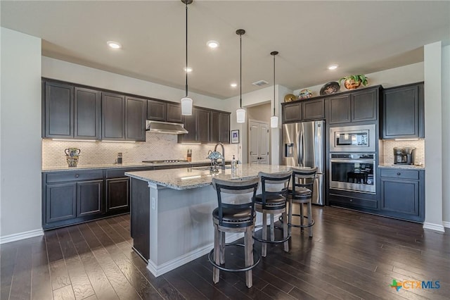 kitchen featuring dark wood finished floors, a center island with sink, stainless steel appliances, light stone countertops, and under cabinet range hood