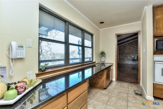 kitchen with light tile patterned floors, stainless steel microwave, ornamental molding, oven, and dark stone counters