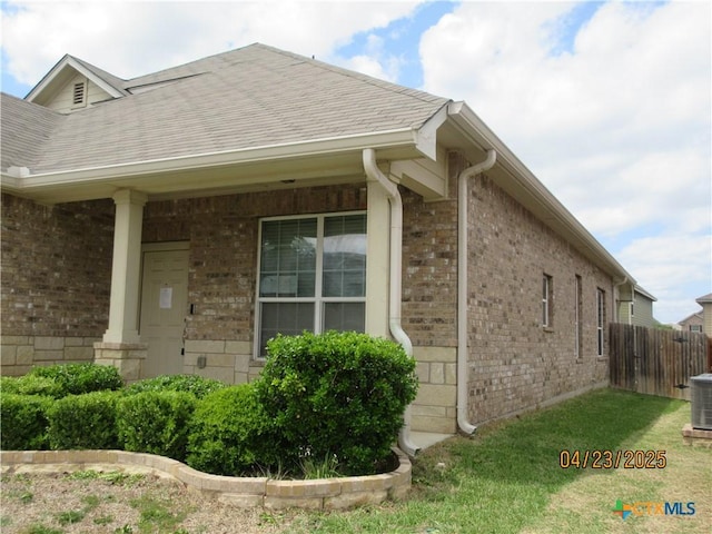 view of side of property with brick siding, fence, central AC unit, and roof with shingles