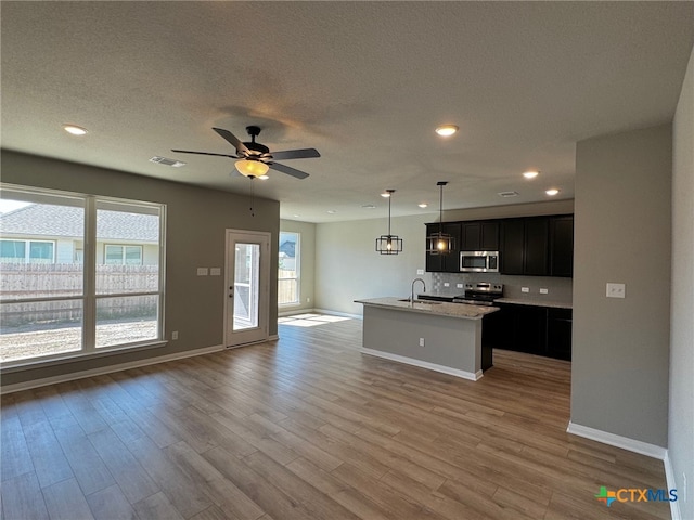 kitchen with light hardwood / wood-style floors, appliances with stainless steel finishes, ceiling fan, an island with sink, and hanging light fixtures