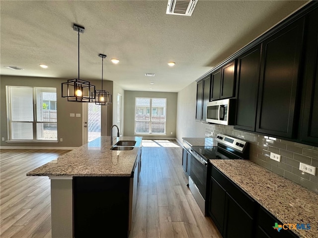 kitchen with sink, a kitchen island with sink, light wood-type flooring, appliances with stainless steel finishes, and decorative light fixtures