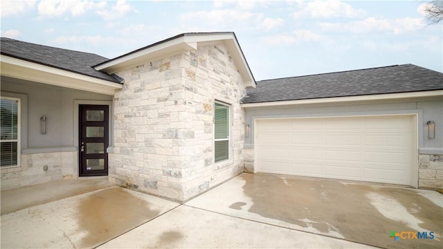 doorway to property with stone siding, a garage, driveway, and a shingled roof