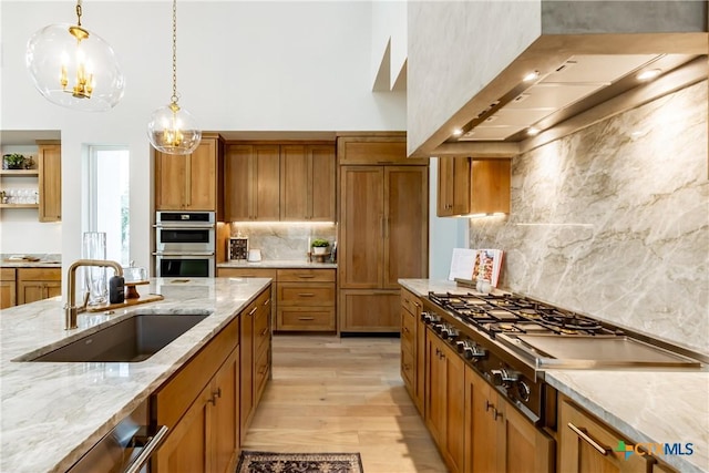 kitchen featuring pendant lighting, stainless steel appliances, brown cabinetry, a sink, and wall chimney exhaust hood