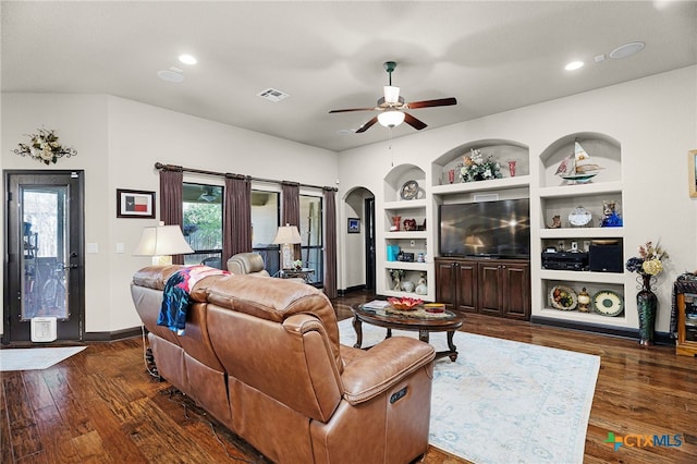 living room with built in shelves, dark wood-style flooring, baseboards, visible vents, and a ceiling fan