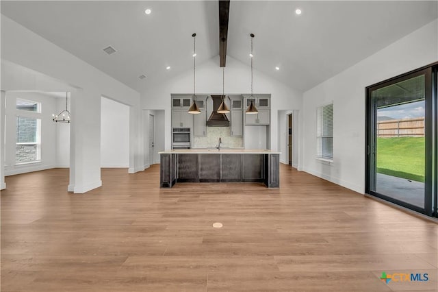 kitchen featuring custom exhaust hood, light wood-type flooring, an island with sink, beam ceiling, and a chandelier