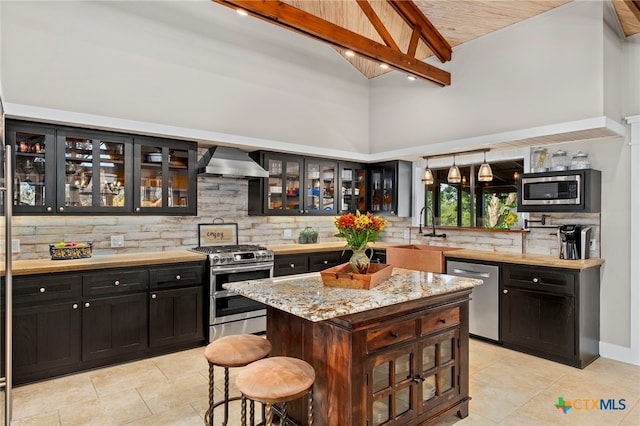 kitchen featuring high vaulted ceiling, tasteful backsplash, wall chimney range hood, appliances with stainless steel finishes, and a sink