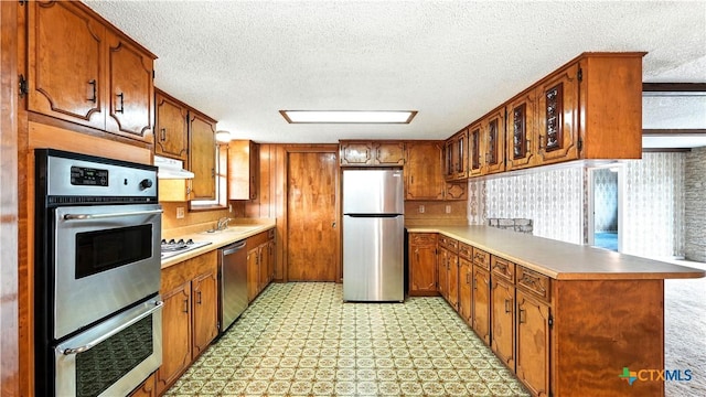 162 Lone Oak St, Seguin, TX 78155 - kitchen with appliances with stainless steel finishes, a peninsula, a textured ceiling, brown cabinetry, and under cabinet range hood