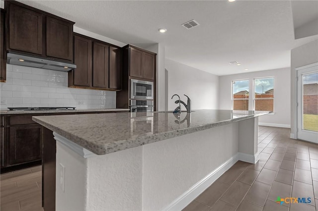 kitchen with light stone counters, sink, decorative backsplash, and a kitchen island with sink