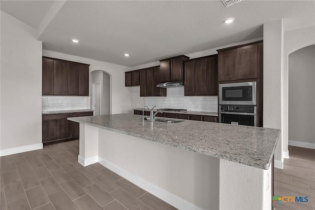 kitchen featuring decorative backsplash, appliances with stainless steel finishes, dark brown cabinets, sink, and an island with sink