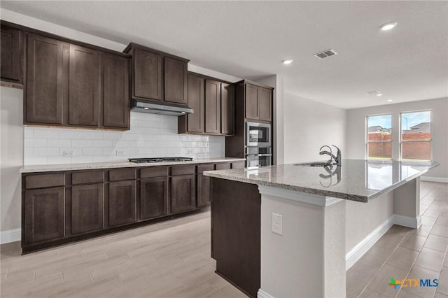 kitchen with a kitchen island with sink, sink, light stone countertops, appliances with stainless steel finishes, and dark brown cabinets