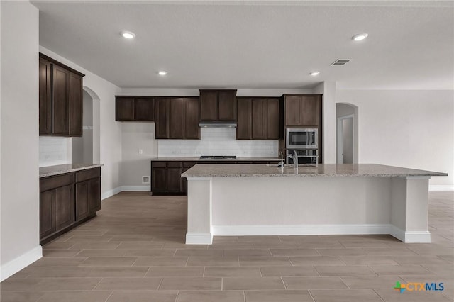 kitchen featuring dark brown cabinetry, stainless steel appliances, light stone counters, and a kitchen island with sink