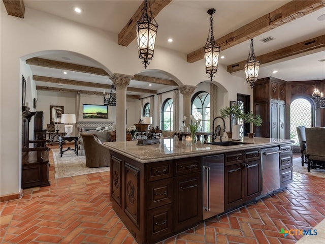 kitchen featuring dark brown cabinetry, sink, decorative light fixtures, a kitchen island with sink, and decorative columns