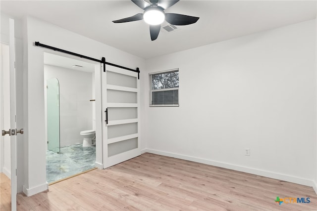 unfurnished bedroom featuring ceiling fan, a barn door, ensuite bath, and light wood-type flooring