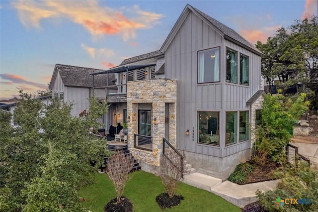 modern farmhouse style home with a balcony, stone siding, roof with shingles, a front lawn, and board and batten siding