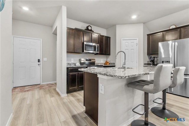 kitchen with a kitchen breakfast bar, light hardwood / wood-style flooring, stainless steel appliances, light stone counters, and a center island with sink