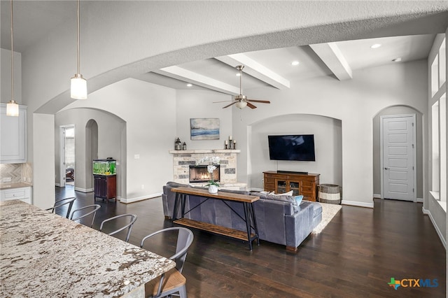 living area with beam ceiling, dark wood-type flooring, a ceiling fan, a stone fireplace, and baseboards