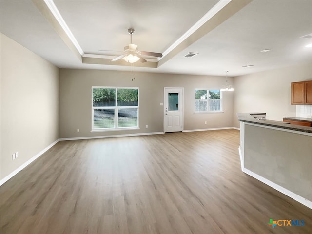 unfurnished living room featuring ceiling fan with notable chandelier, light wood-type flooring, and a raised ceiling