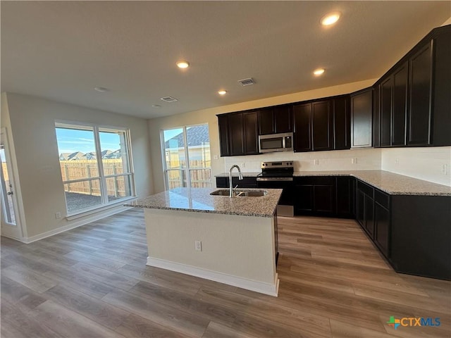 kitchen with appliances with stainless steel finishes, sink, a kitchen island with sink, light stone counters, and light hardwood / wood-style floors