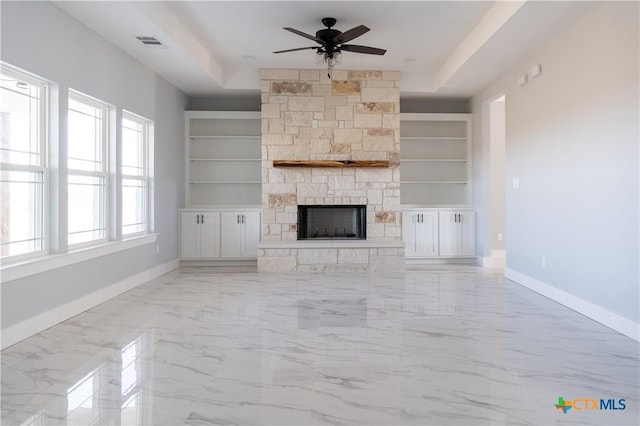 unfurnished living room with ceiling fan, a stone fireplace, a raised ceiling, and built in features