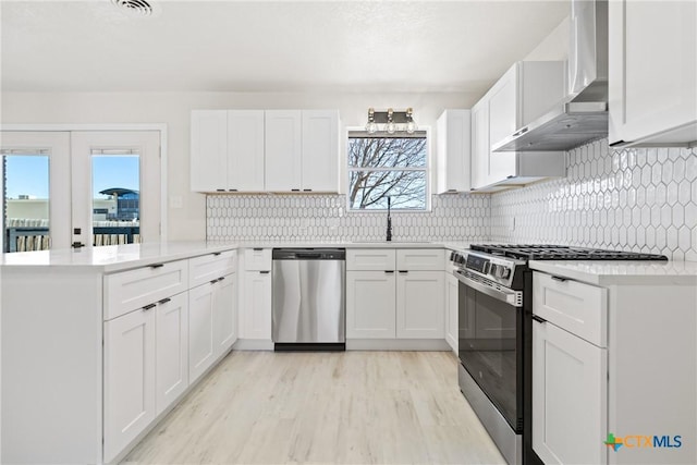 kitchen with light countertops, appliances with stainless steel finishes, white cabinets, wall chimney range hood, and a peninsula