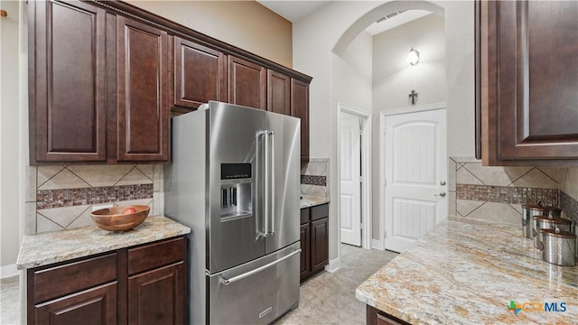 kitchen featuring high end fridge, decorative backsplash, dark brown cabinets, arched walkways, and light stone counters