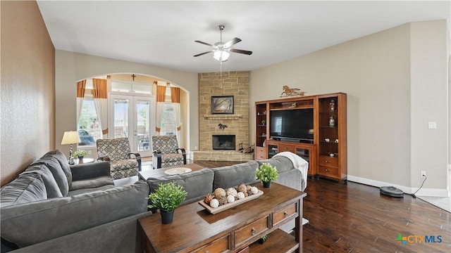 living area with arched walkways, a ceiling fan, dark wood-style flooring, baseboards, and a stone fireplace