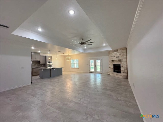 unfurnished living room featuring a stone fireplace, a tray ceiling, ornamental molding, and ceiling fan with notable chandelier