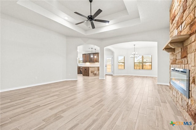 unfurnished living room featuring a stone fireplace, ceiling fan with notable chandelier, a raised ceiling, and light wood-type flooring