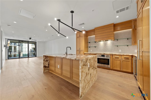 kitchen with an island with sink, sink, decorative backsplash, stainless steel oven, and light stone countertops