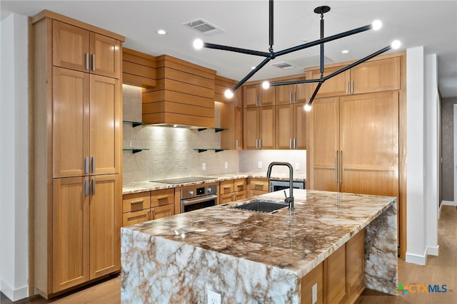 kitchen with light stone countertops, black electric cooktop, decorative light fixtures, oven, and a large island with sink