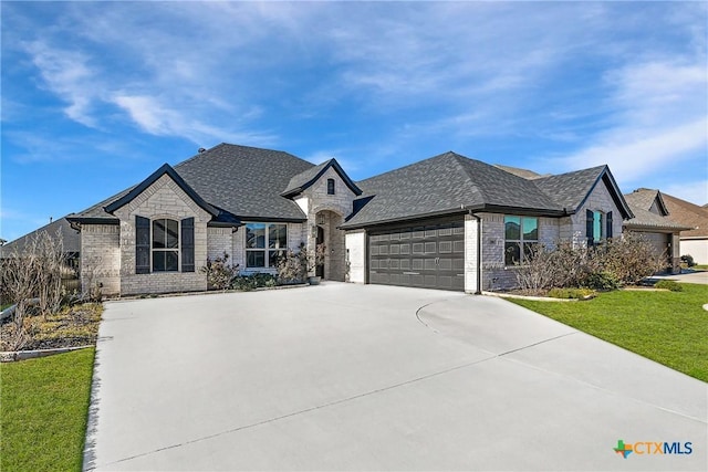 french country style house with driveway, a front yard, a shingled roof, a garage, and brick siding