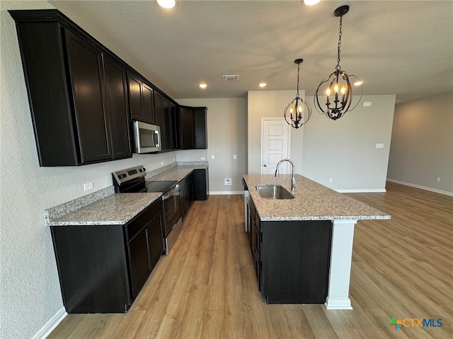 kitchen with light stone counters, sink, an island with sink, light wood-type flooring, and appliances with stainless steel finishes