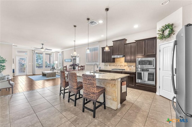 kitchen featuring appliances with stainless steel finishes, an island with sink, sink, hanging light fixtures, and light stone counters