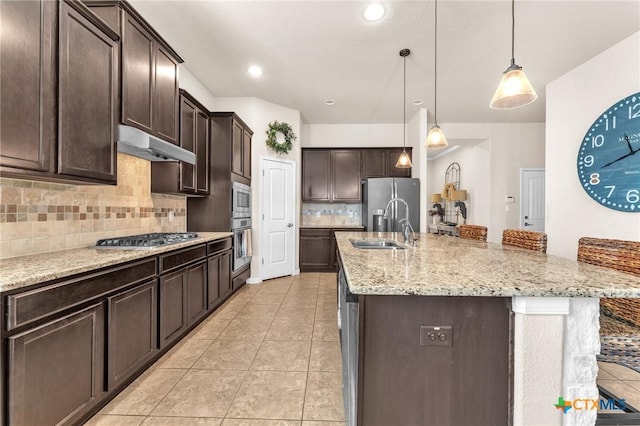 kitchen with appliances with stainless steel finishes, hanging light fixtures, light stone counters, dark brown cabinets, and a center island with sink