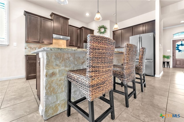 kitchen featuring pendant lighting, stainless steel fridge, dark brown cabinets, a kitchen breakfast bar, and light stone countertops