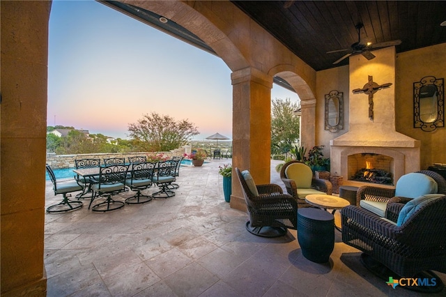 patio terrace at dusk with a ceiling fan, outdoor dining space, and a fireplace