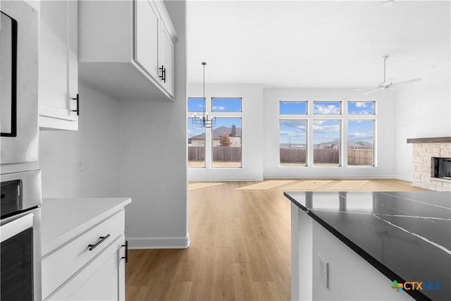 kitchen with light wood finished floors, white cabinets, oven, a stone fireplace, and ceiling fan with notable chandelier