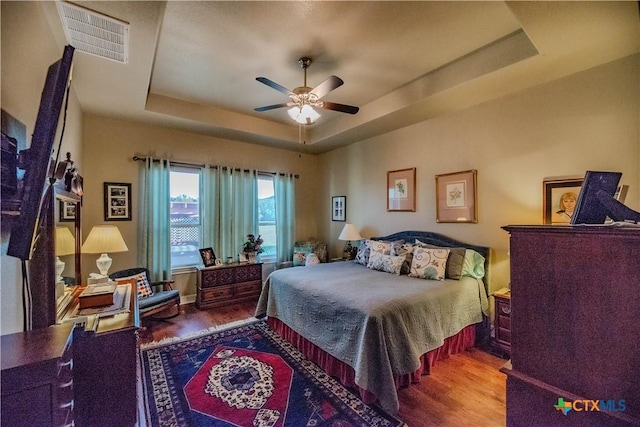 bedroom with ceiling fan, a tray ceiling, wood finished floors, and visible vents