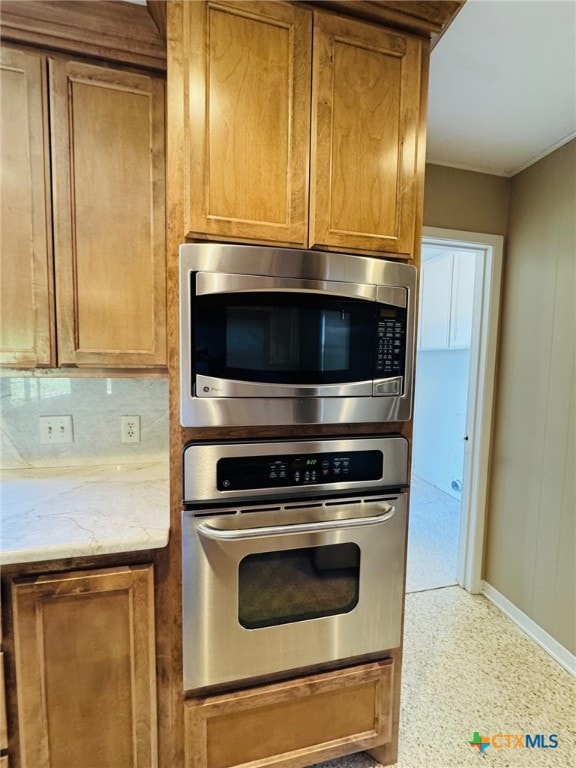 kitchen featuring baseboards, appliances with stainless steel finishes, brown cabinets, backsplash, and light stone counters