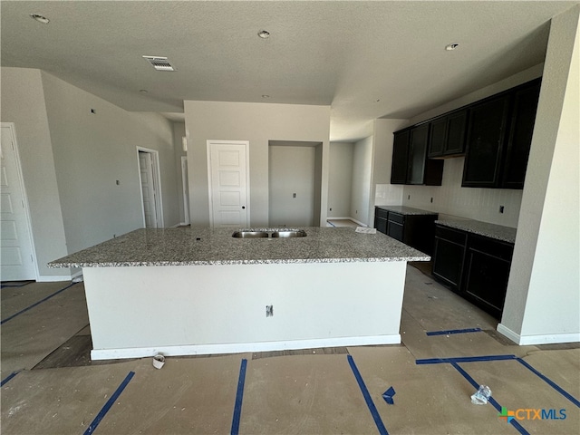 kitchen with concrete floors, sink, light stone countertops, a textured ceiling, and a kitchen island with sink