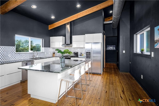 kitchen featuring white cabinets, wall chimney exhaust hood, beamed ceiling, a kitchen island, and stainless steel appliances