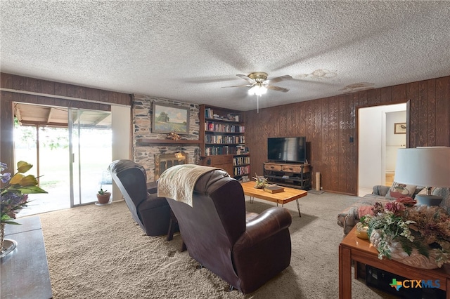 living room featuring a textured ceiling, carpet, a ceiling fan, wooden walls, and a stone fireplace