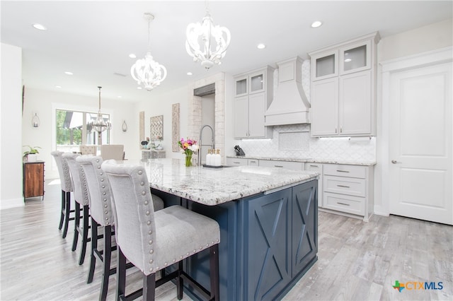 kitchen featuring a center island with sink, tasteful backsplash, light hardwood / wood-style flooring, decorative light fixtures, and premium range hood