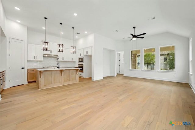 kitchen featuring white cabinetry, tasteful backsplash, decorative light fixtures, light wood-type flooring, and an island with sink