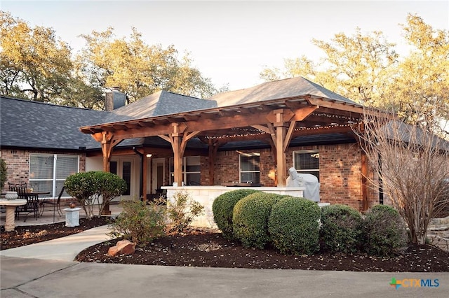 view of front facade featuring a patio, roof with shingles, a pergola, a chimney, and brick siding