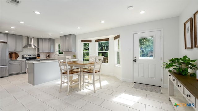 kitchen with wall chimney exhaust hood, light tile patterned floors, gray cabinets, a kitchen island, and appliances with stainless steel finishes