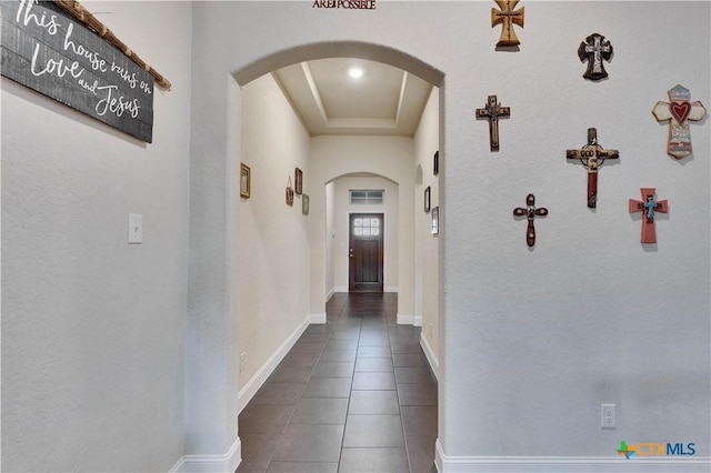 hallway featuring arched walkways, a raised ceiling, tile patterned flooring, and baseboards